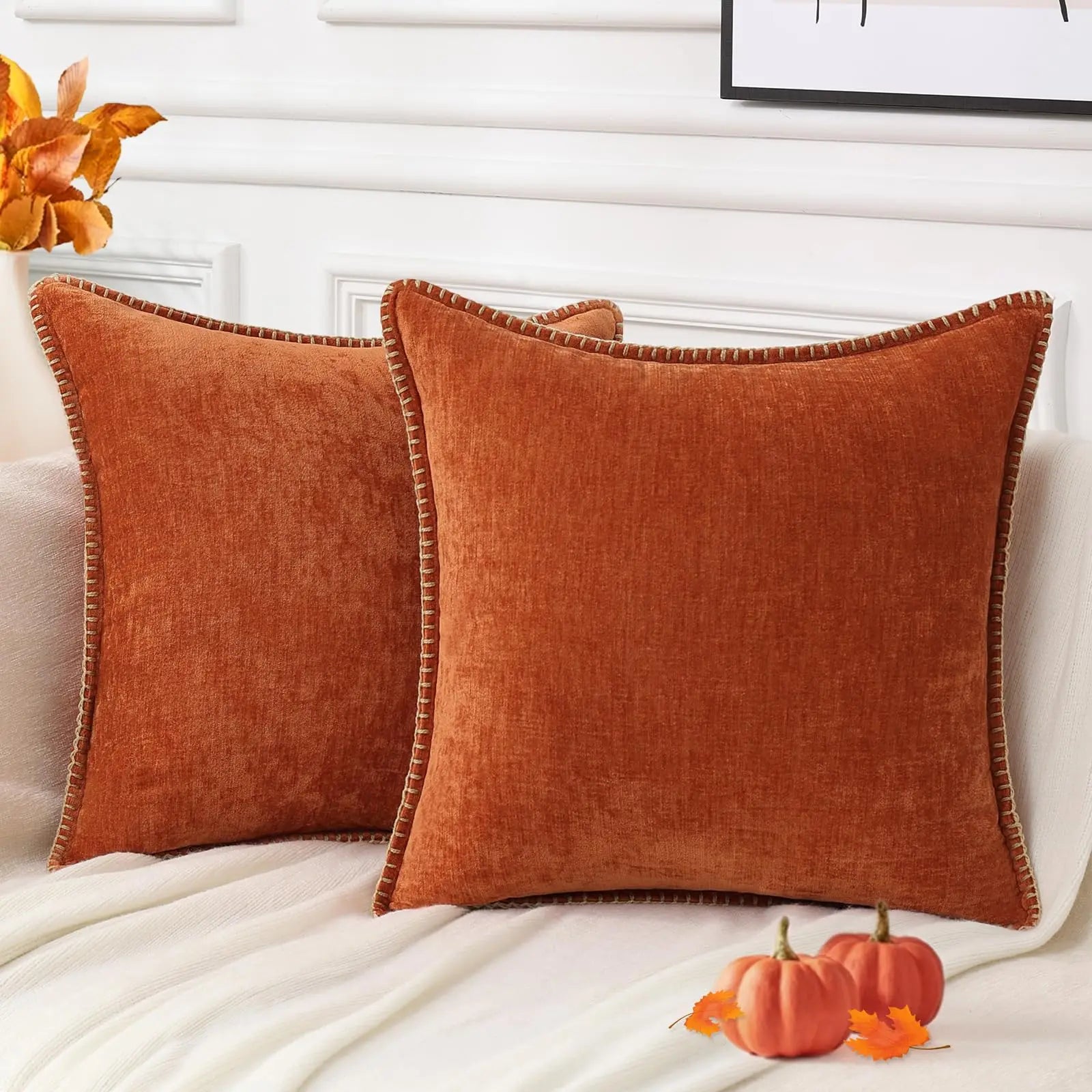 Two orange pillows on a bed with pumpkins and flowers in the background