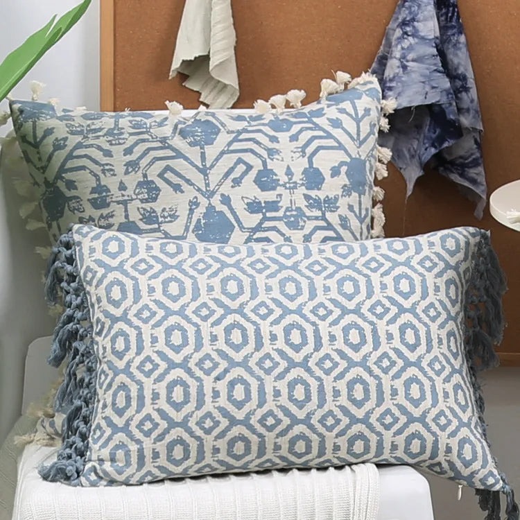 Two blue patterned pillows with tassels on a white surface, with a wooden headboard and blanket in the background.