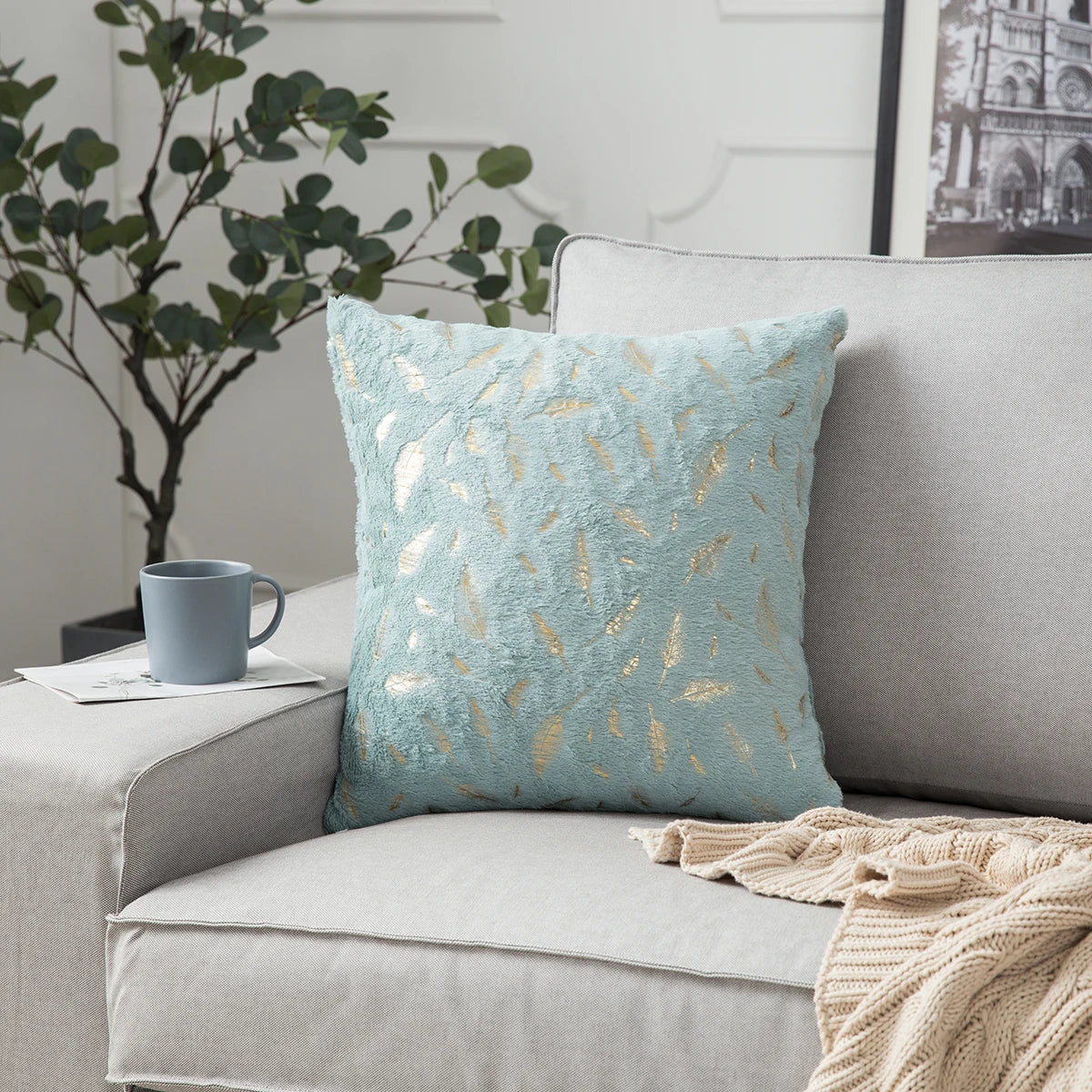 Light blue pillow with gold feather pattern on a gray sofa with a plant and mug in the background.