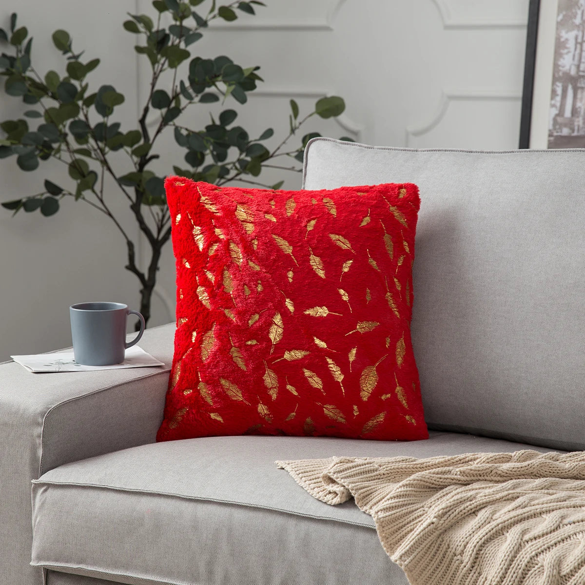 Red pillow with gold leaf pattern on a gray sofa with a plant and cup in the background