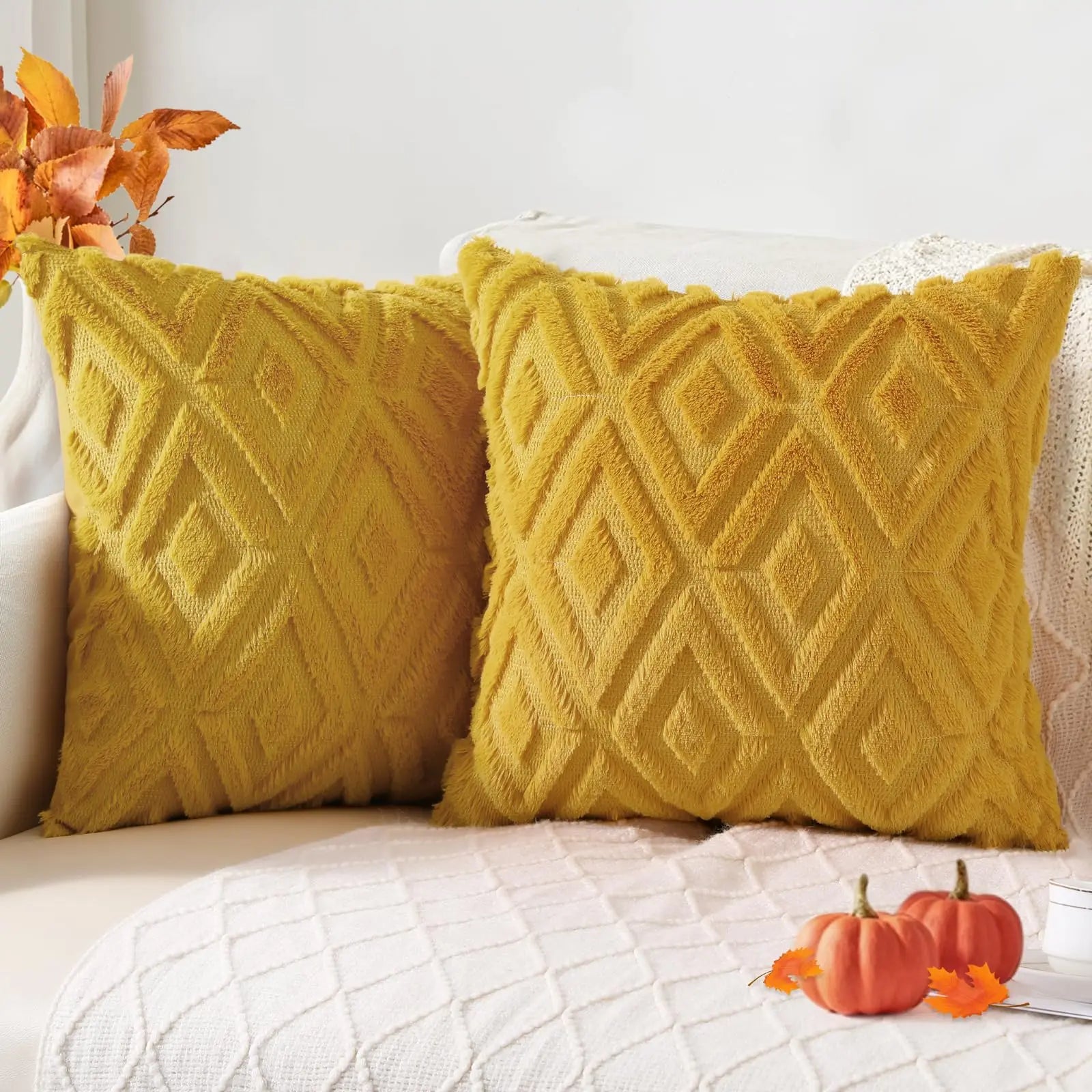 Two yellow textured pillows on a white couch with small pumpkins and autumn leaves.